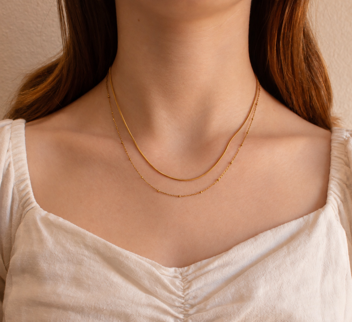 A close-up of a woman's neck and upper chest wearing a delicate double-layered gold chain necklace over a cream-colored top.