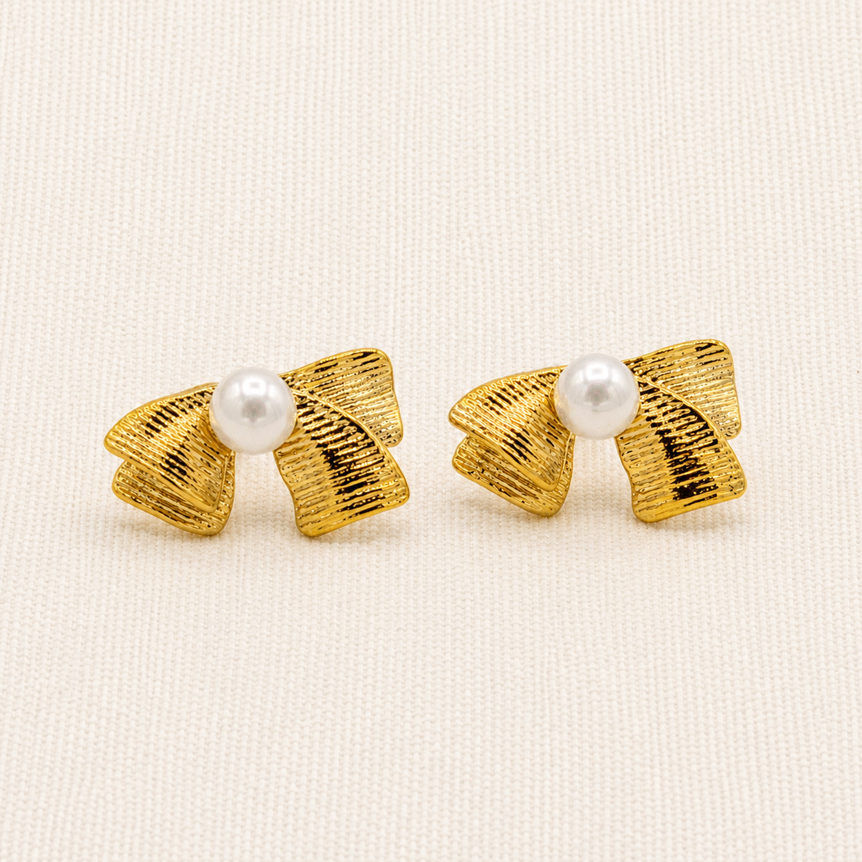A pair of gold bow-shaped stud earrings with a textured ribbed finish and a small white pearl at the center