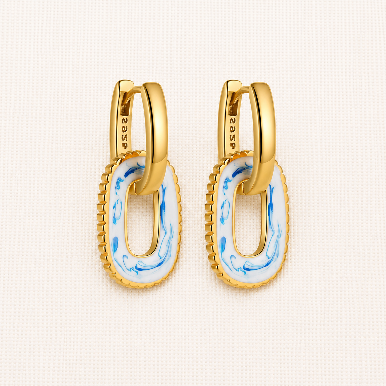 A pair of gold huggie hoop earrings with oval drop charms featuring a white enamel center with blue marbled swirls and a textured gold border. 
